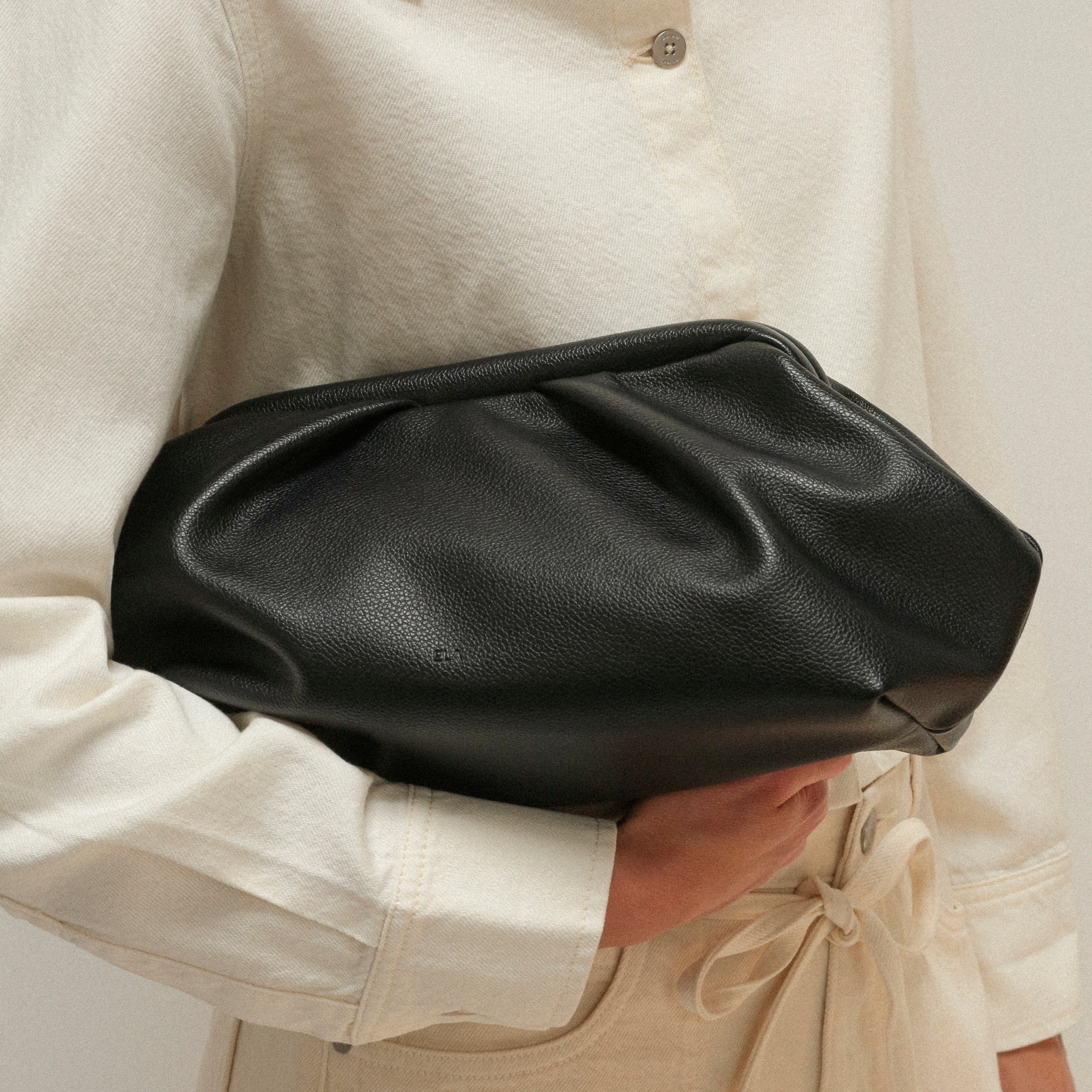 A person in a cream-colored shirt and tie-dye trousers holds a black pebbled vegan leather clutch bag